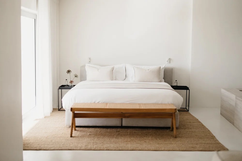 A bed on a jute rug under a high white wall, a bench at the foot of it